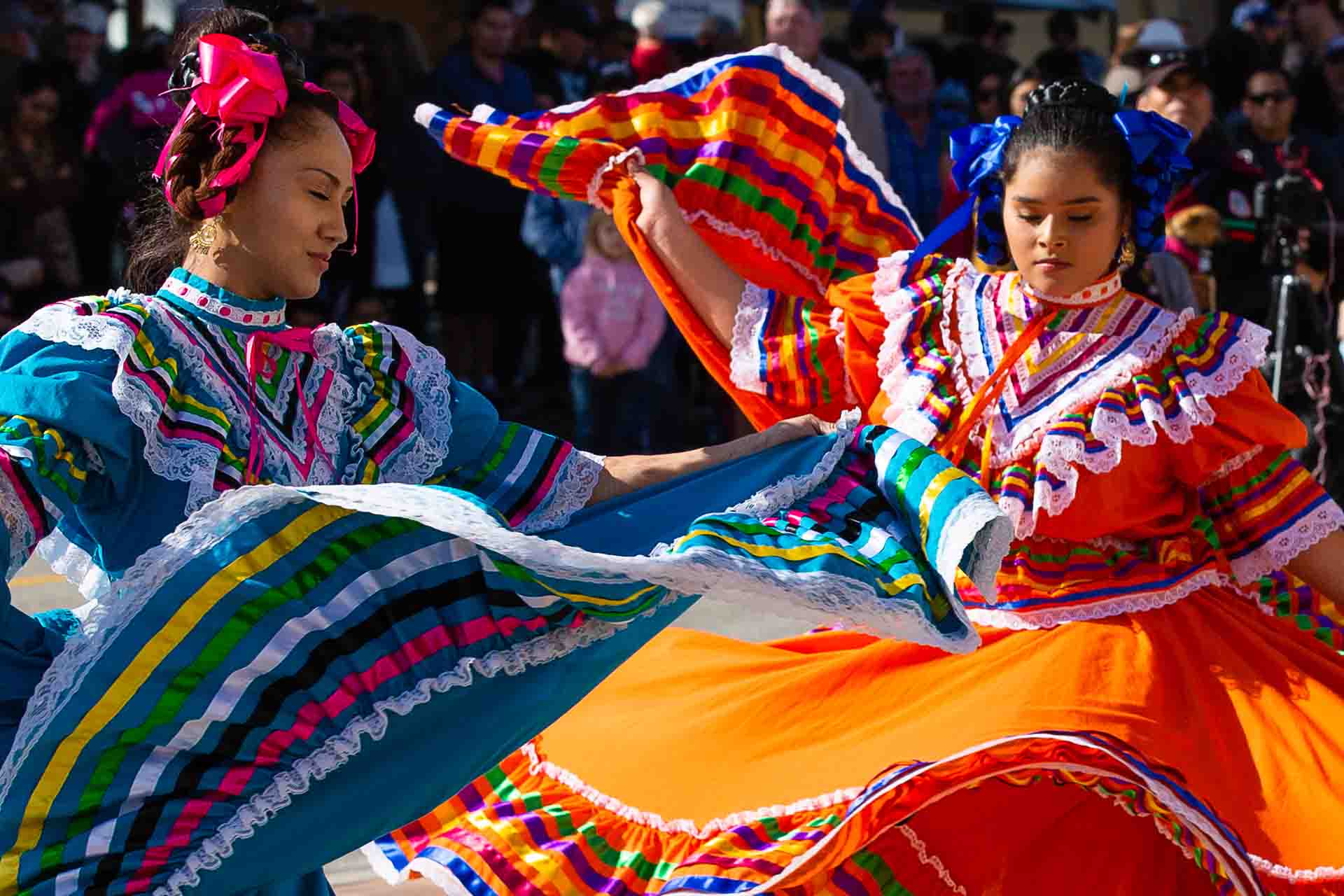 Image of two folkloric dancers in bright colored dresses, with full flowing skirts rippling as they dance.