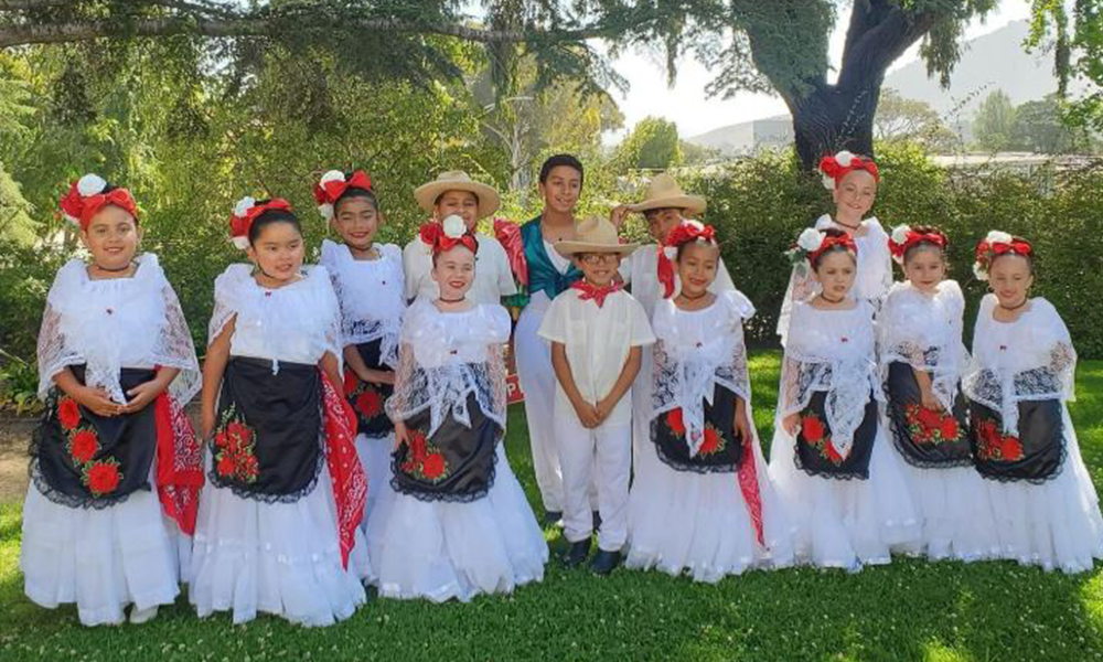 Group of children smiling and posing in a shaded, green outdoor setting, wearing traditional red, white, and black folklórico attire.