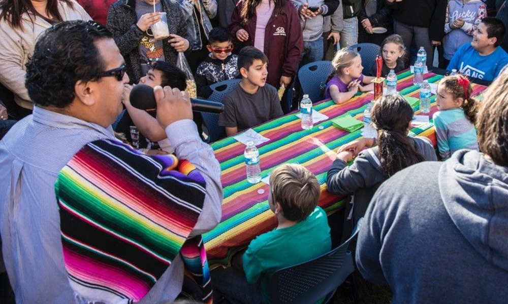 Image of kids at a long table preparing for a Tamale Eating Contest, with the event Mc and crowd standing by. - Photo by Rick Evans